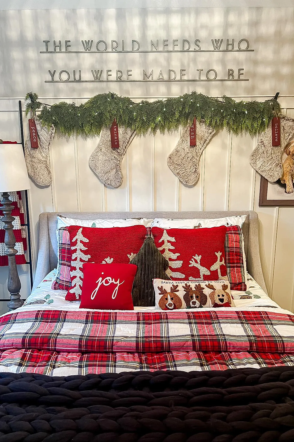 Christmas bedroom featuring a beige upholstered bed with red plaid bedding and pillows, including red pillows with white tree and reindeer silhouettes, a small red pillow with 'joy' script, green textured tree-shaped pillow, a decorative pillow with reindeer faces reading 'Santa paws is coming to town', four fluffy cream stockings hanging above the headboard on greenery garland with tags reading peace, twinkle, wish, believe.