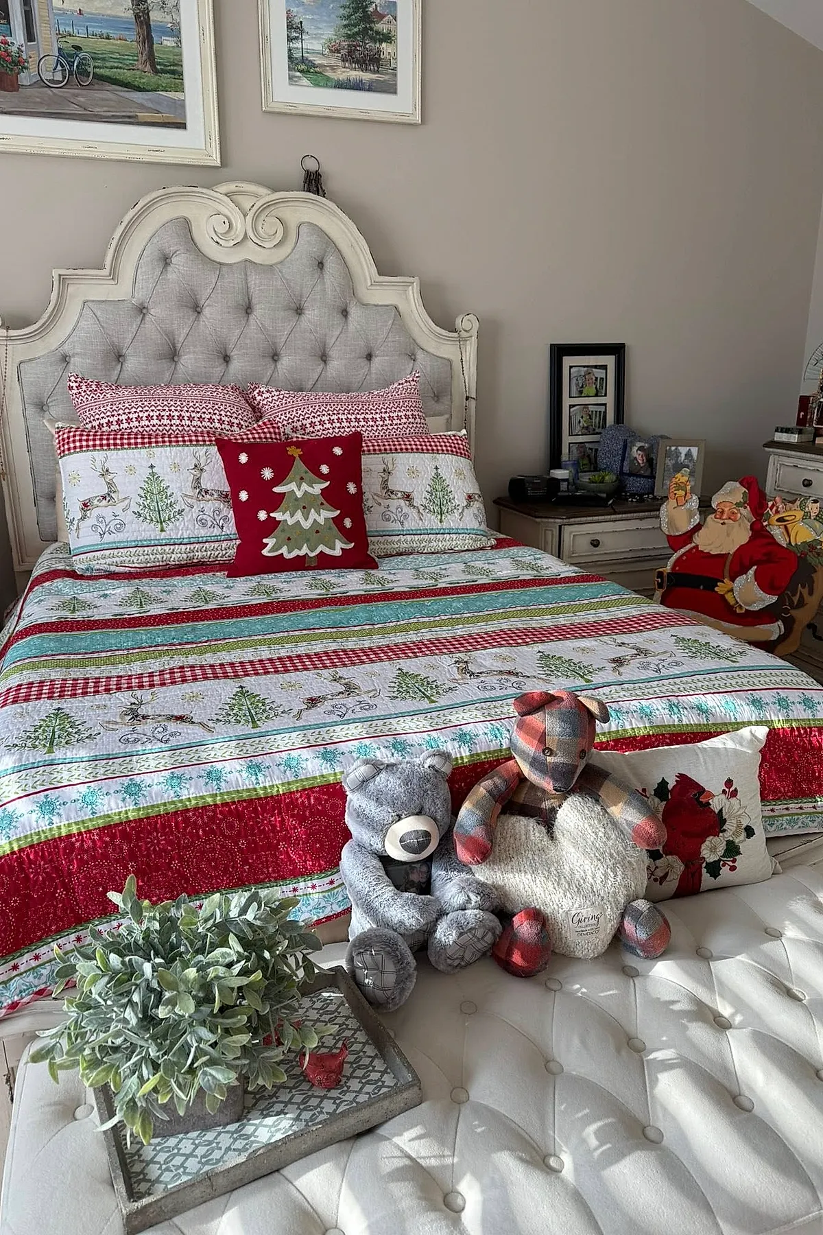 Christmas bedroom featuring a tufted upholstered headboard, bedding with red, green, and white stripes decorated with reindeer and Christmas trees, a red pillow with an embroidered tree in the center, two stuffed teddy bears on a white tufted bench at the foot of the bed alongside a pillow with cardinals and greenery. A vintage Santa figure is visible near the bedside table.