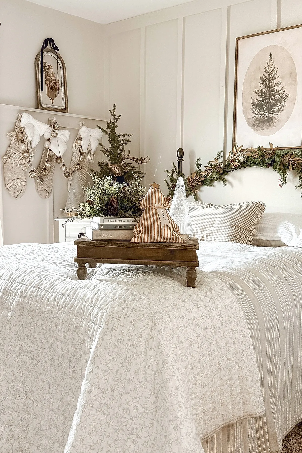 Neutral-toned Christmas bedroom featuring a white quilted bedspread, striped bedding, gingham pillows, a wooden tray on the bed holding books and fabric Christmas tree decorations, wall-mounted greenery garland above the headboard, four textured cream stockings hanging on the wall, small decorated pine trees and rustic holiday decor throughout.