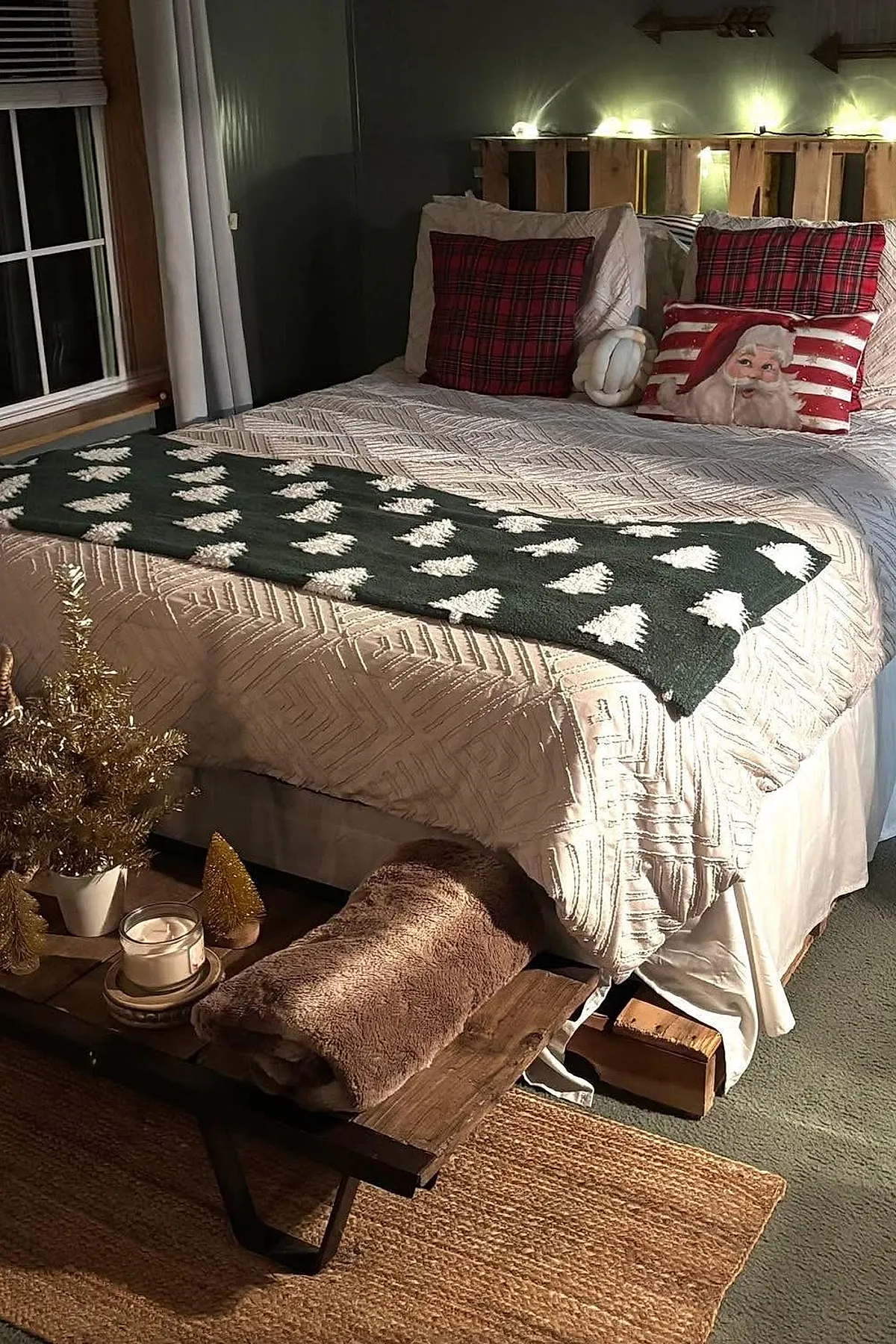 Christmas bedroom with a wooden pallet headboard adorned with warm string lights, beige patterned bedding, two red plaid pillows, a Santa Claus pillow with stripes, a dark green throw blanket with white Christmas tree patterns, a rustic wooden bench at the foot of the bed holding gold miniature Christmas trees, a candle in a glass holder, and a folded brown fuzzy blanket.