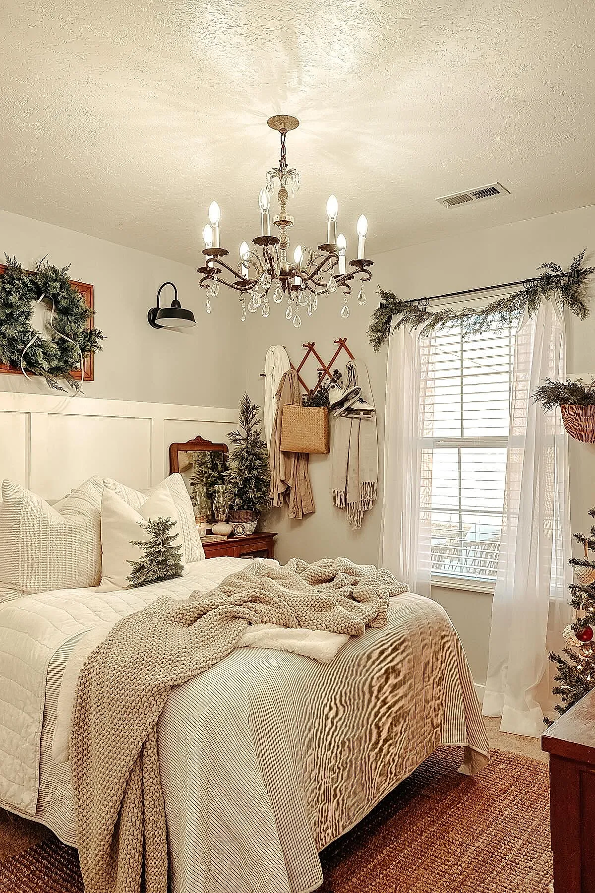 Christmas bedroom decorated in neutral tones featuring a bed with cream and beige bedding, a chunky knit blanket draped over the edge, pillows with pine tree prints, a small Christmas tree on the bedside table, green garlands over the window and wreath on the wall, accented by soft natural light.