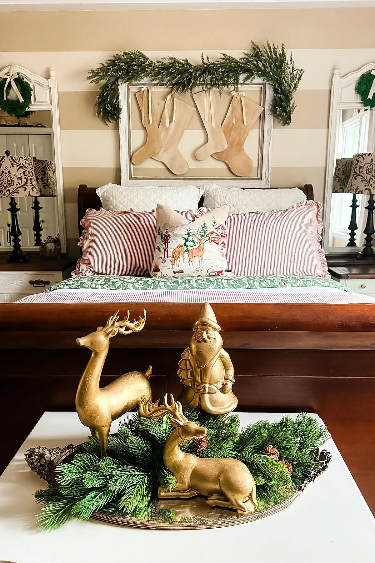 Christmas bedroom featuring a wooden sleigh bed with red and white striped pillows, white quilted shams, a decorative pillow with reindeer print, framed wooden stockings above the headboard decorated with pine garland, two ornate mirrors with wreaths on either side, and a white table in front displaying gold figurines of two reindeer and Santa surrounded by pine branches.
