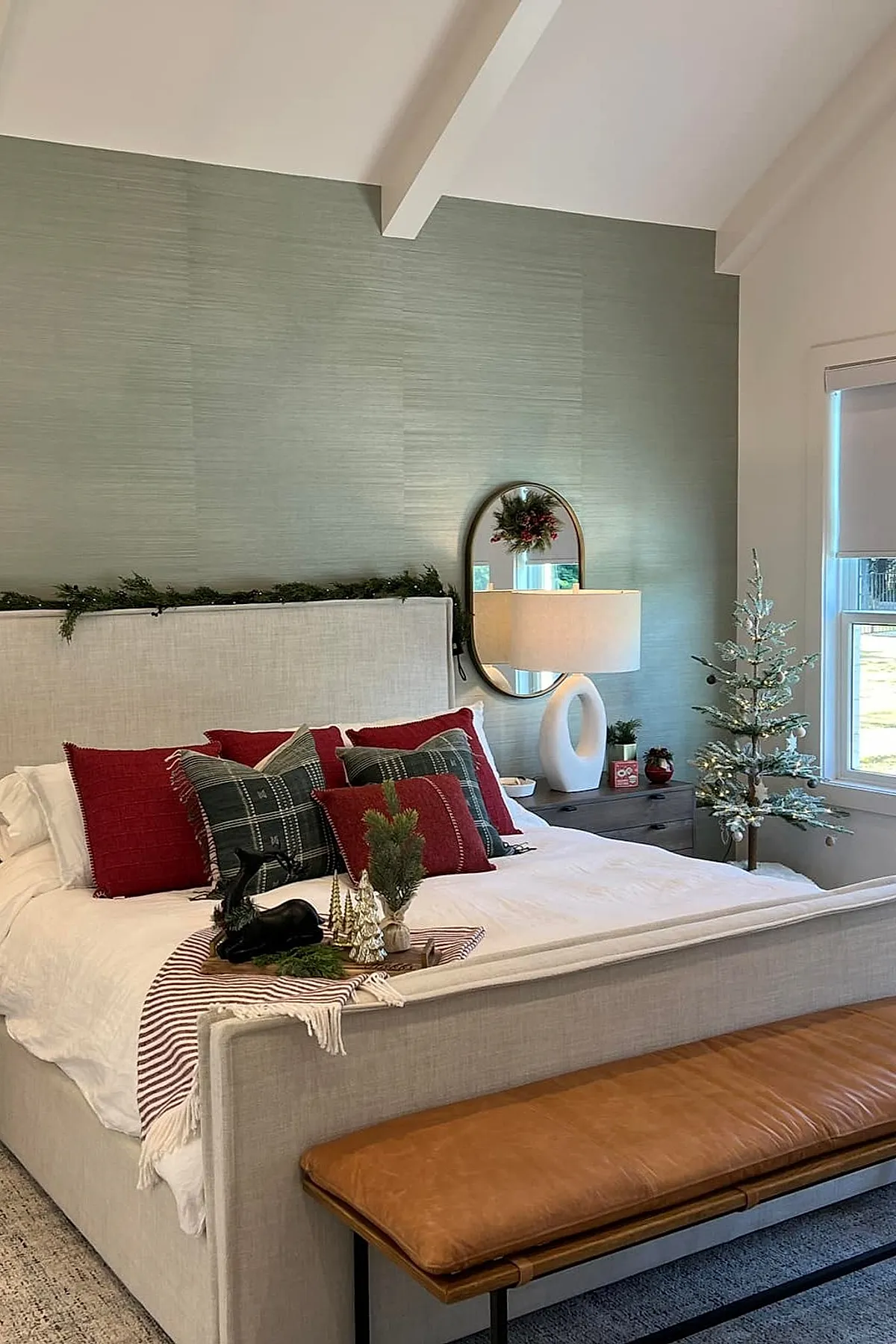 Christmas bedroom featuring a beige upholstered bed with red and green plaid pillows, solid red pillows, white bedding, a small decorated Christmas tree next to the window, greenery garland along the headboard, a round mirror with wreath reflection above the nightstand holding a modern white lamp and holiday decor.
