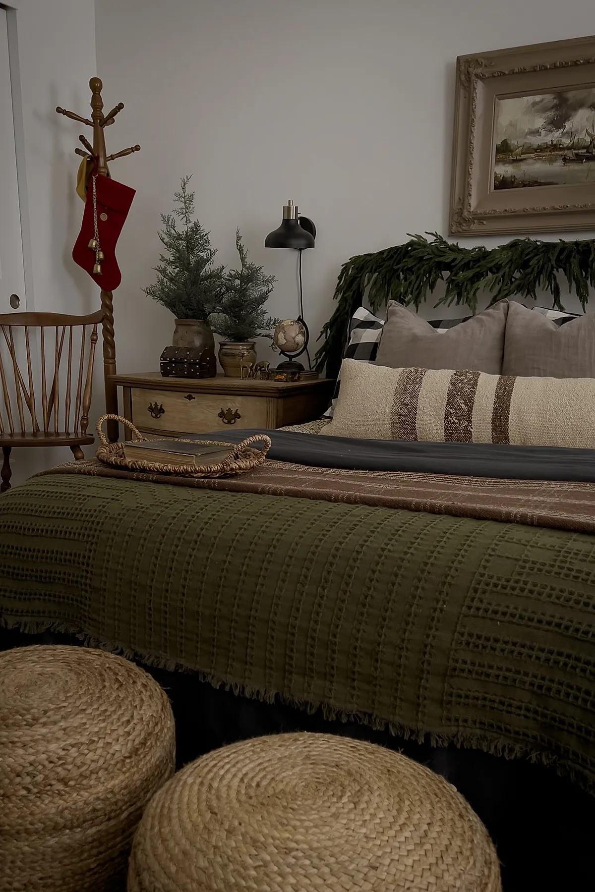 Christmas bedroom featuring an olive green textured blanket on the bed, beige and brown striped lumbar pillow, gray and buffalo check pillows, a garland of greenery draped along the headboard, wooden side table with potted small pine trees and rustic decor, wooden chair with red Christmas stocking hanging from a coat rack.