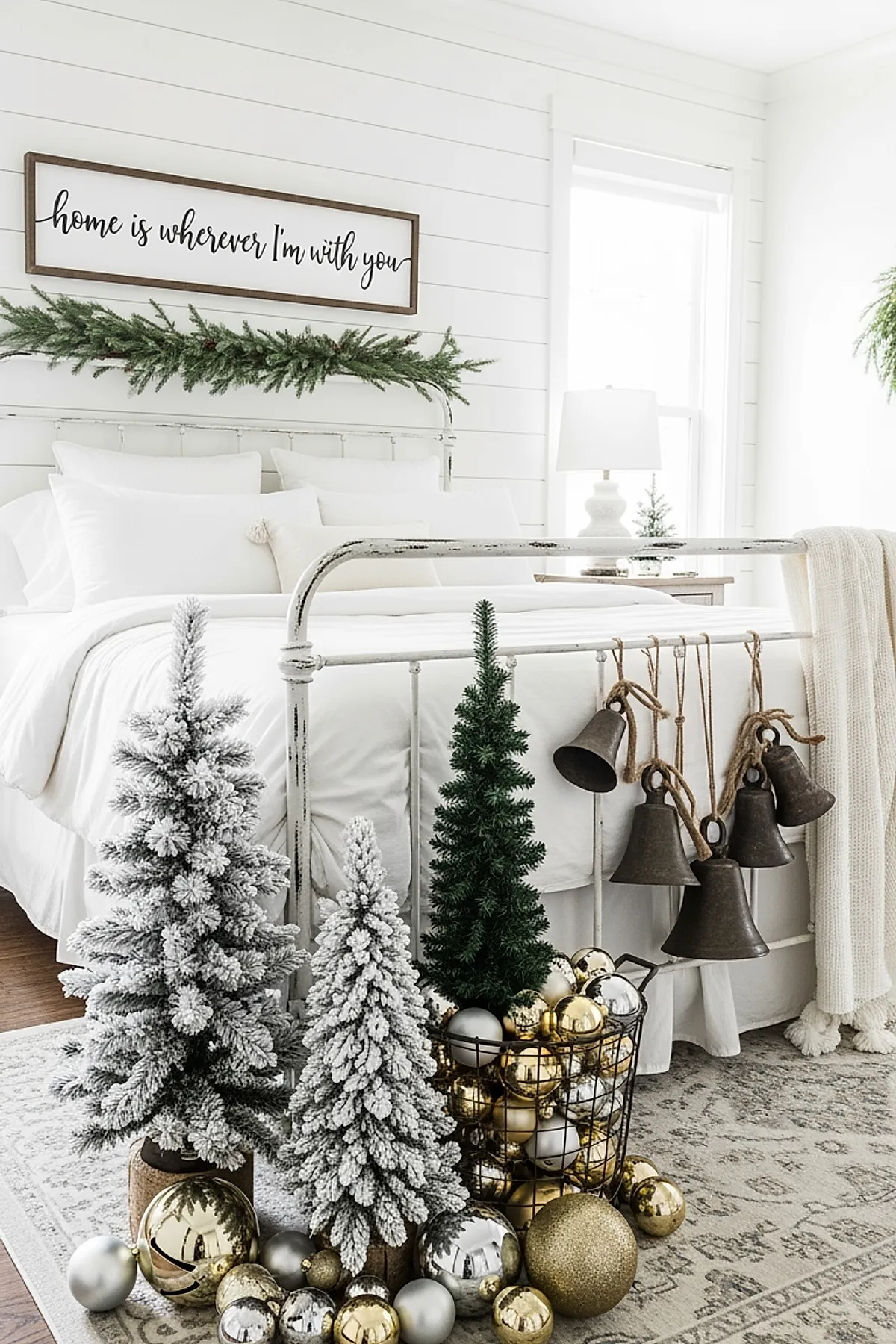 Bright white Christmas bedroom featuring an iron bed with white bedding, frosted mini Christmas trees in front of the bed, gold and silver ornaments scattered on a patterned gray rug, and five large rustic metal bells hanging from the footboard.