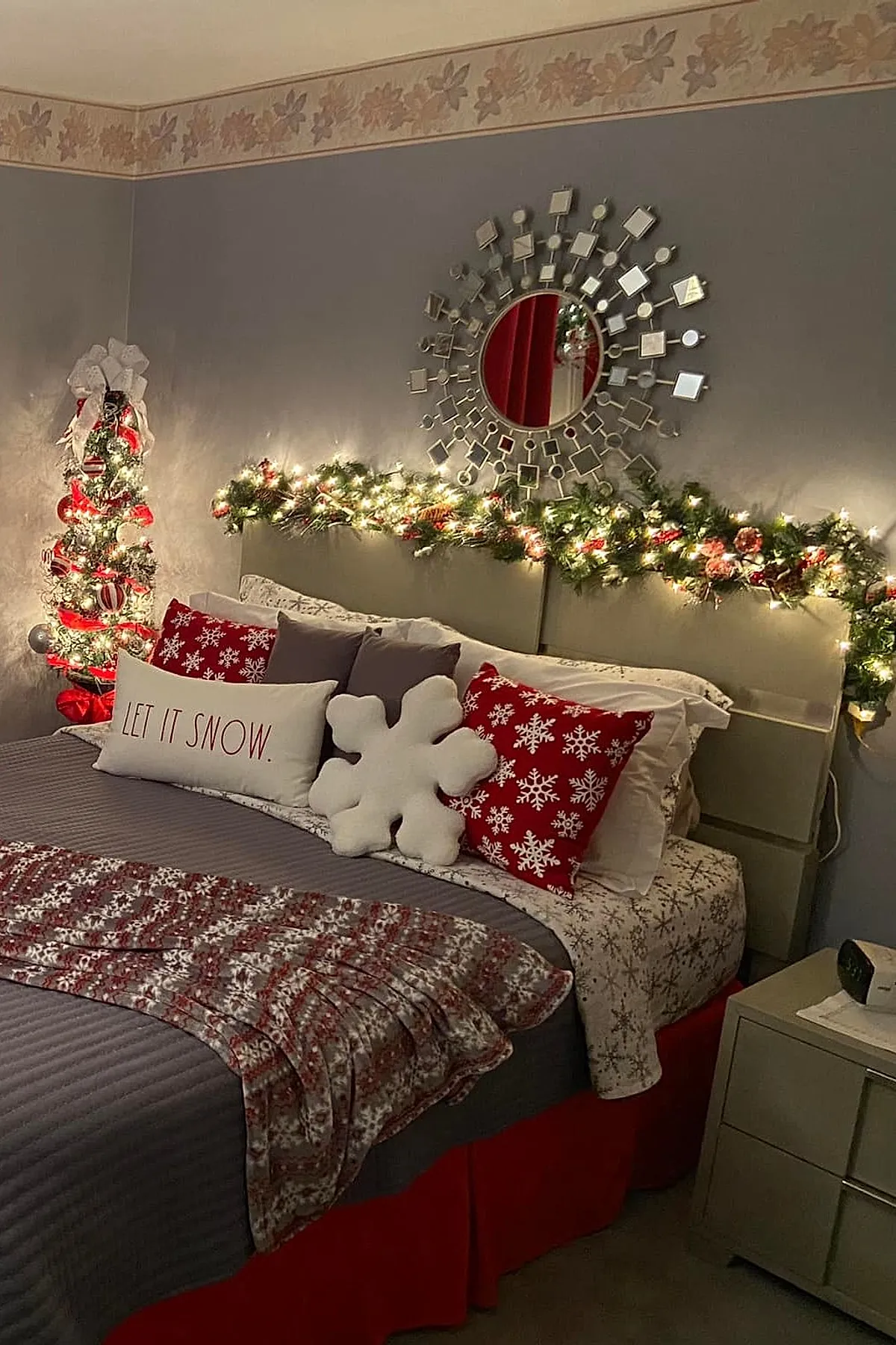 Christmas bedroom with a gray upholstered headboard decorated with green garland wrapped in warm white string lights, bed layered with red and white snowflake print pillows, a white pillow with 'LET IT SNOW' text, and a large white snowflake-shaped cushion. A small decorated Christmas tree adorned with red ribbons and ornaments stands in the corner against gray walls.
