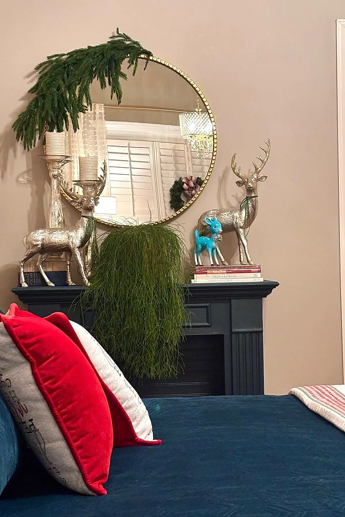 Christmas bedroom featuring two large gold reindeer figurines, one on each side of a black mantel, a small bright blue fawn figurine standing on books on the right side, a round mirror above with greenery draped over the top left edge, tall candle holders with textured white candles on the left, and red and white pillows in the foreground on a dark blue bedspread.