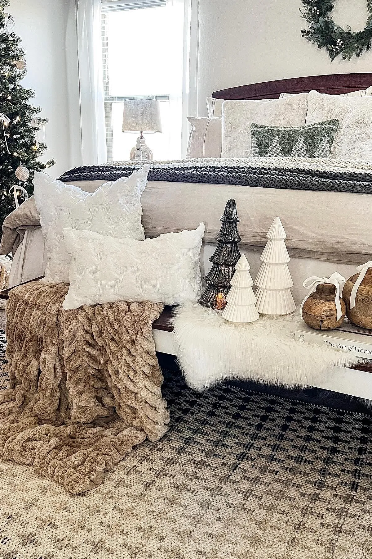 Christmas bedroom corner with a bed dressed in neutral beige bedding, layered with chunky knit gray and cream blankets, accented by white textured pillows. A bench at the foot of the bed features a soft brown plush throw, white faux fur runner, ceramic and metal miniature Christmas trees, wooden ornaments tied with white ribbons, all set against a patterned black and beige rug.