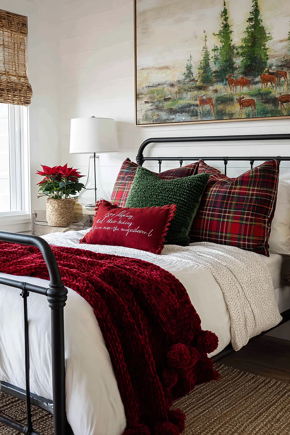 Christmas bedroom featuring a black metal bed frame with white bedding layered with a chunky red knit throw blanket, plaid pillows in red and green tones, a textured dark green pillow, a small red rectangular pillow with white script text, natural woven window shades, a glass lamp with white shade on the bedside table, and rustic wall art depicting deer in a forest scene.