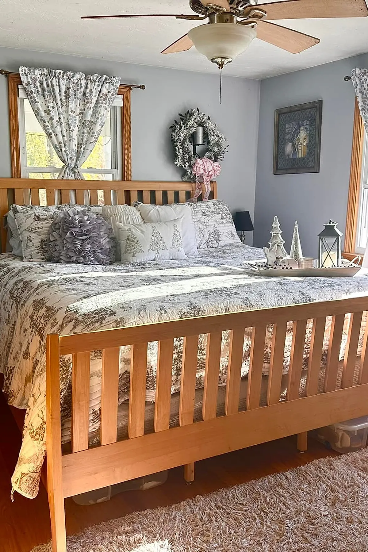 Christmas bedroom with neutral tones featuring a wooden bed frame, gray and white bedding with tree patterns, decorative pillows including a ruffled lavender one and embroidered ones with Christmas trees, a frosted wreath with pink ribbon hanging above the headboard, floral patterned curtains on windows, and a white tray on the bed holding silver and white Christmas tree figurines and a lantern.
