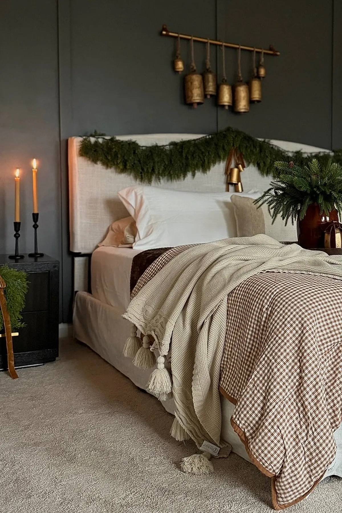 Christmas bedroom with dark gray walls, a beige upholstered headboard decorated with a green garland, beige bedding layered with a cream knit blanket featuring tassels, brass bells hanging above the bed, two lit candles on a black nightstand, and a brown vase filled with evergreen branches.