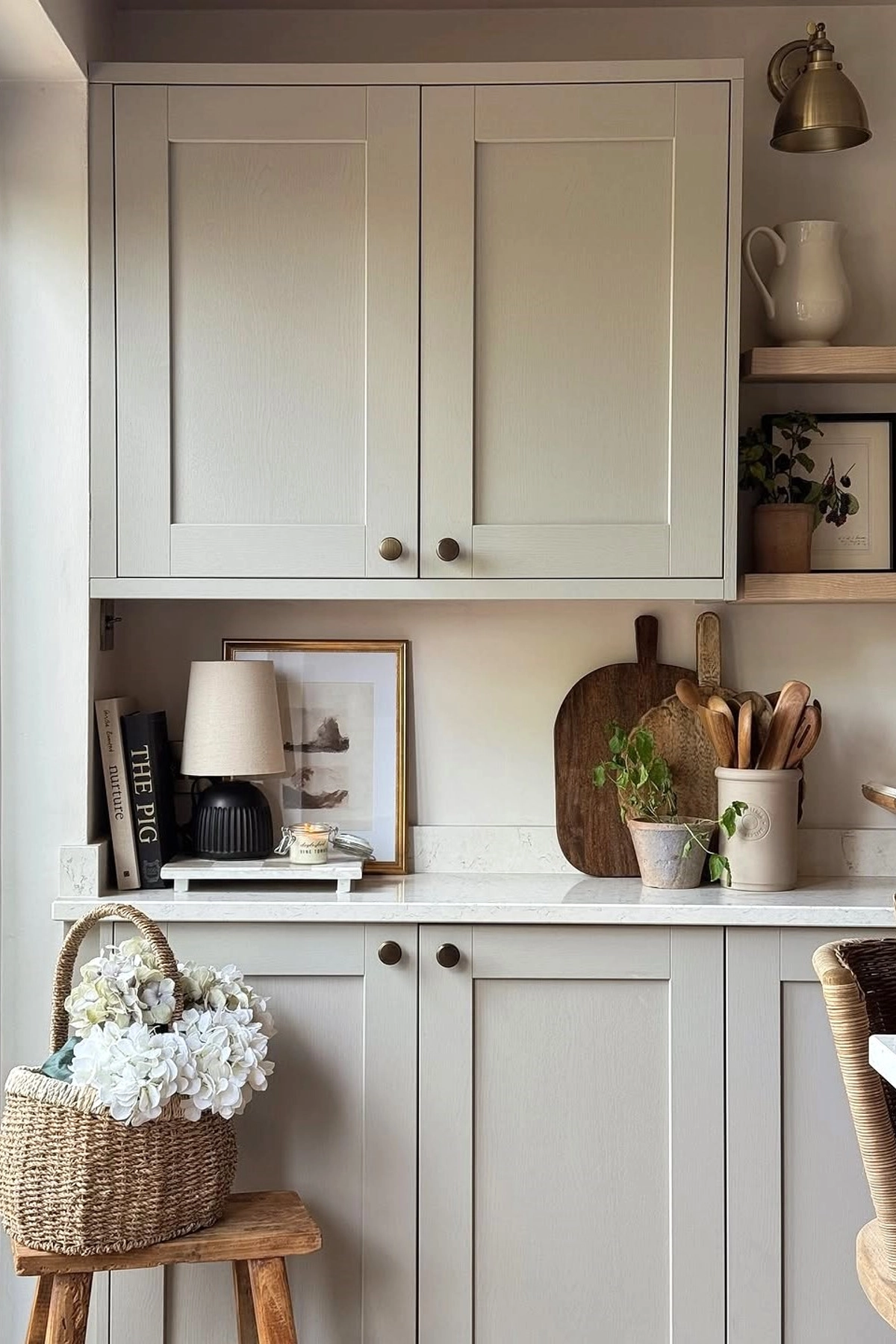 Neutral kitchen with soft gray cabinets, wooden accents, basket with hydrangeas, small lamp, cookbooks, framed picture and potted herbs on the countertop.