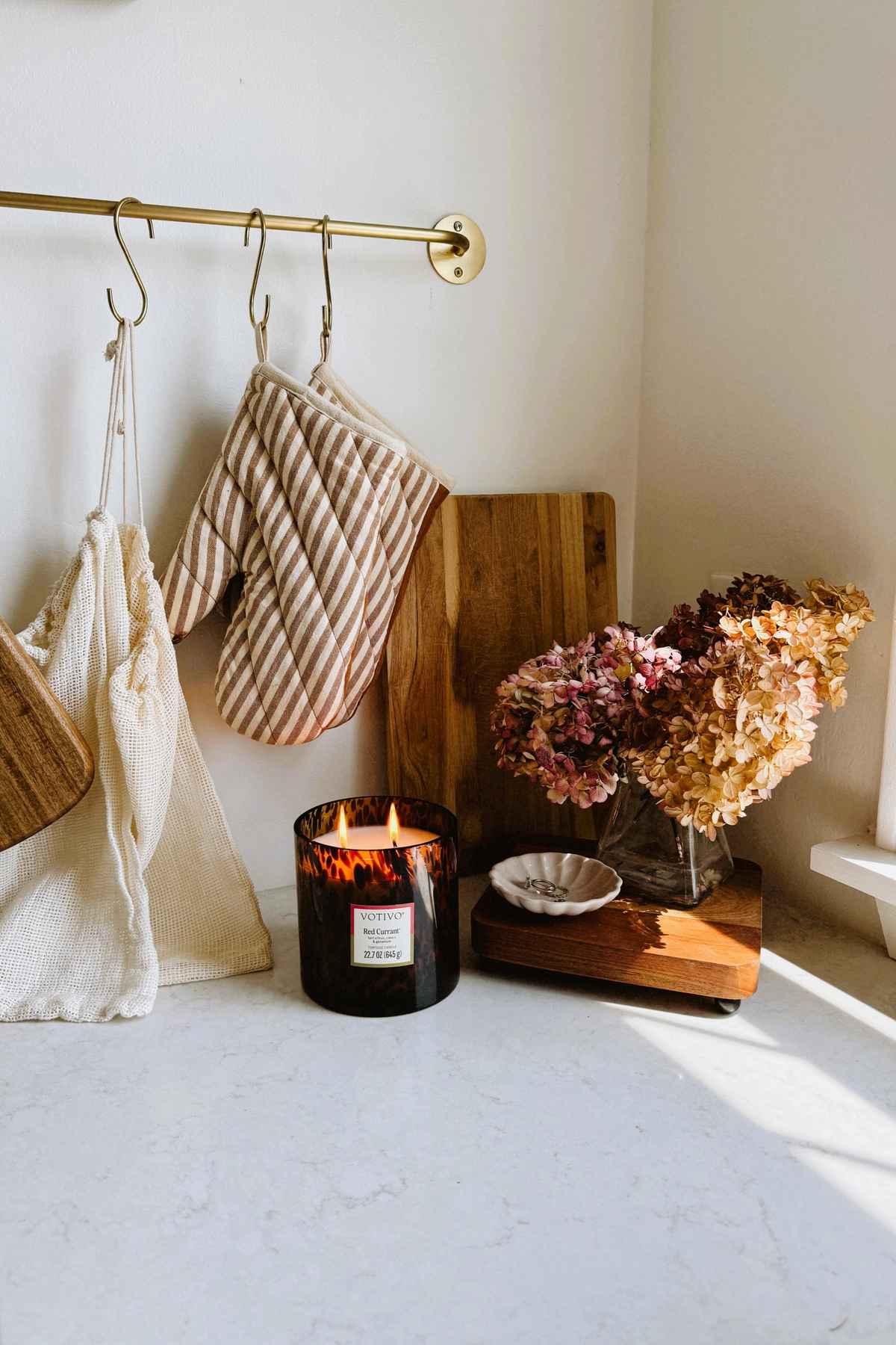 Kitchen countertop decor with striped oven mitts hanging from a brass rail, dried hydrangeas in a glass vase, a lit tortoiseshell candle, wood cutting boards, and a small dish on a white counter.