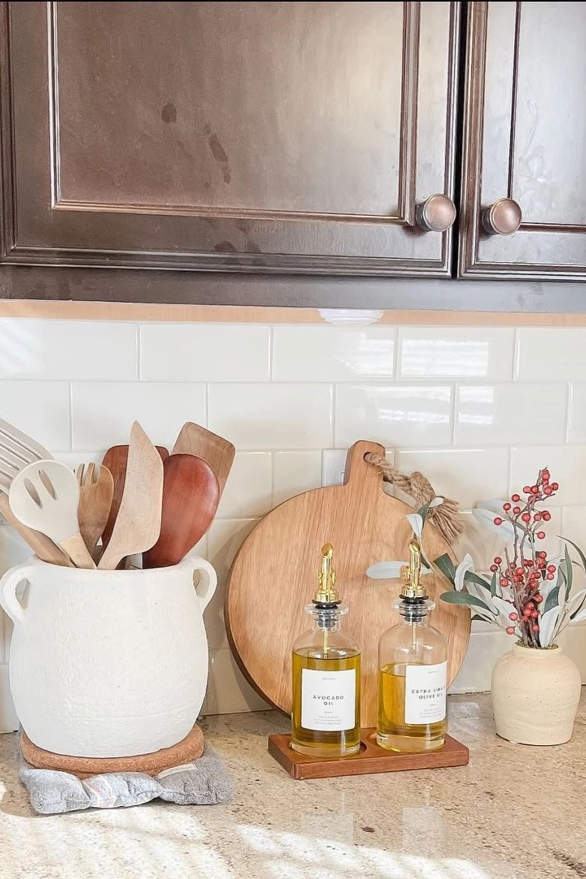 Kitchen countertop with white jar holding wooden utensils, cutting boards, oil bottles, and a small vase with berries.