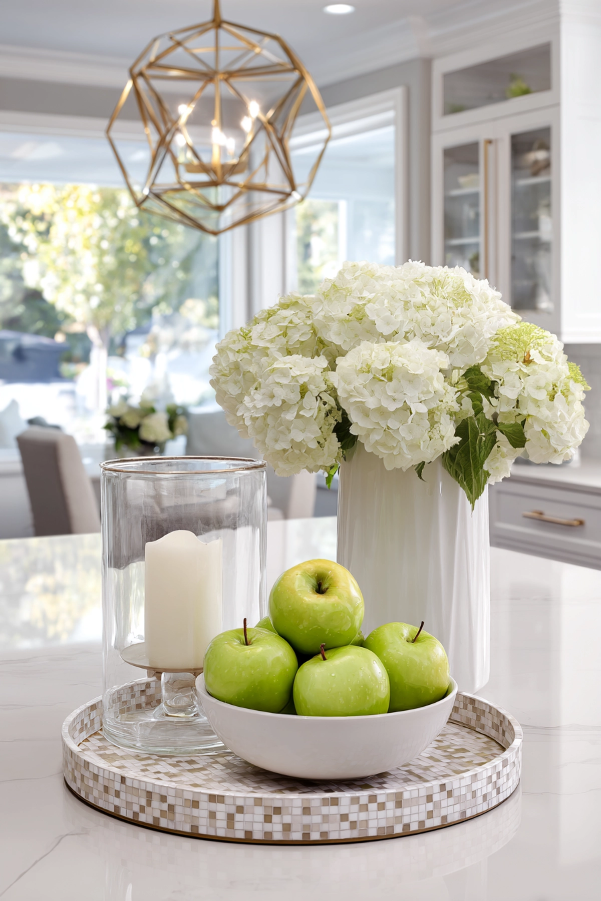 Kitchen island with white hydrangeas in vase, green apples in bowl, candle in glass holder, and geometric gold pendant light overhead.