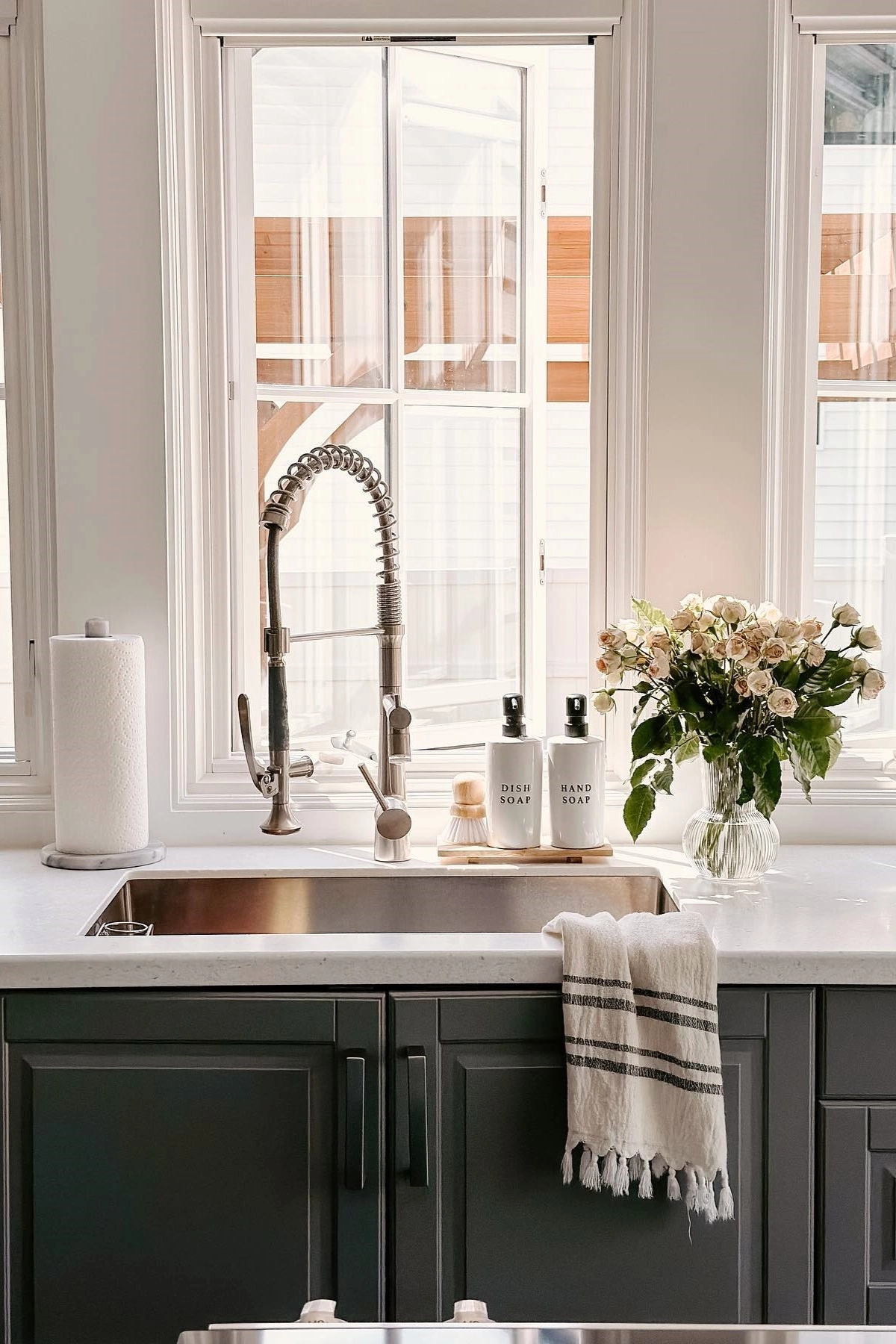 Kitchen sink under large windows with a vase of flowers, hand soap bottles, and a towel on a dark cabinet countertop.