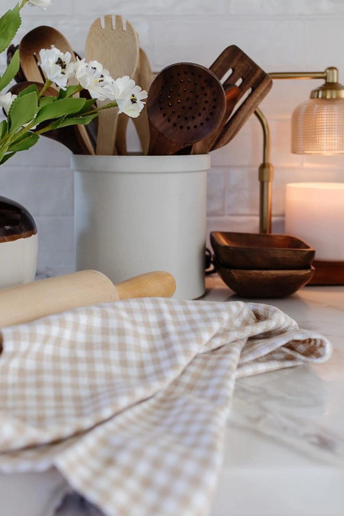 Kitchen countertop with wooden utensils in a white container, a lit candle, vase with flowers, and checked cloth.