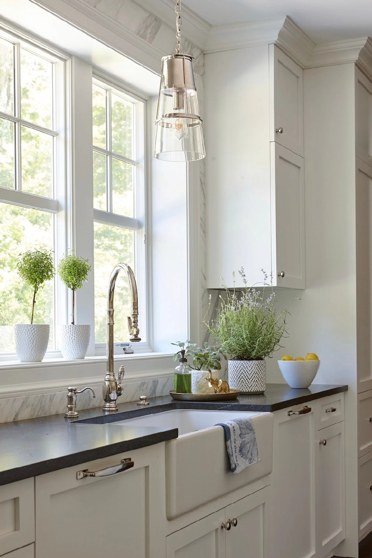 Bright kitchen with white cabinets, silver faucet, plants in pots, and a bowl of lemons on the dark countertop.