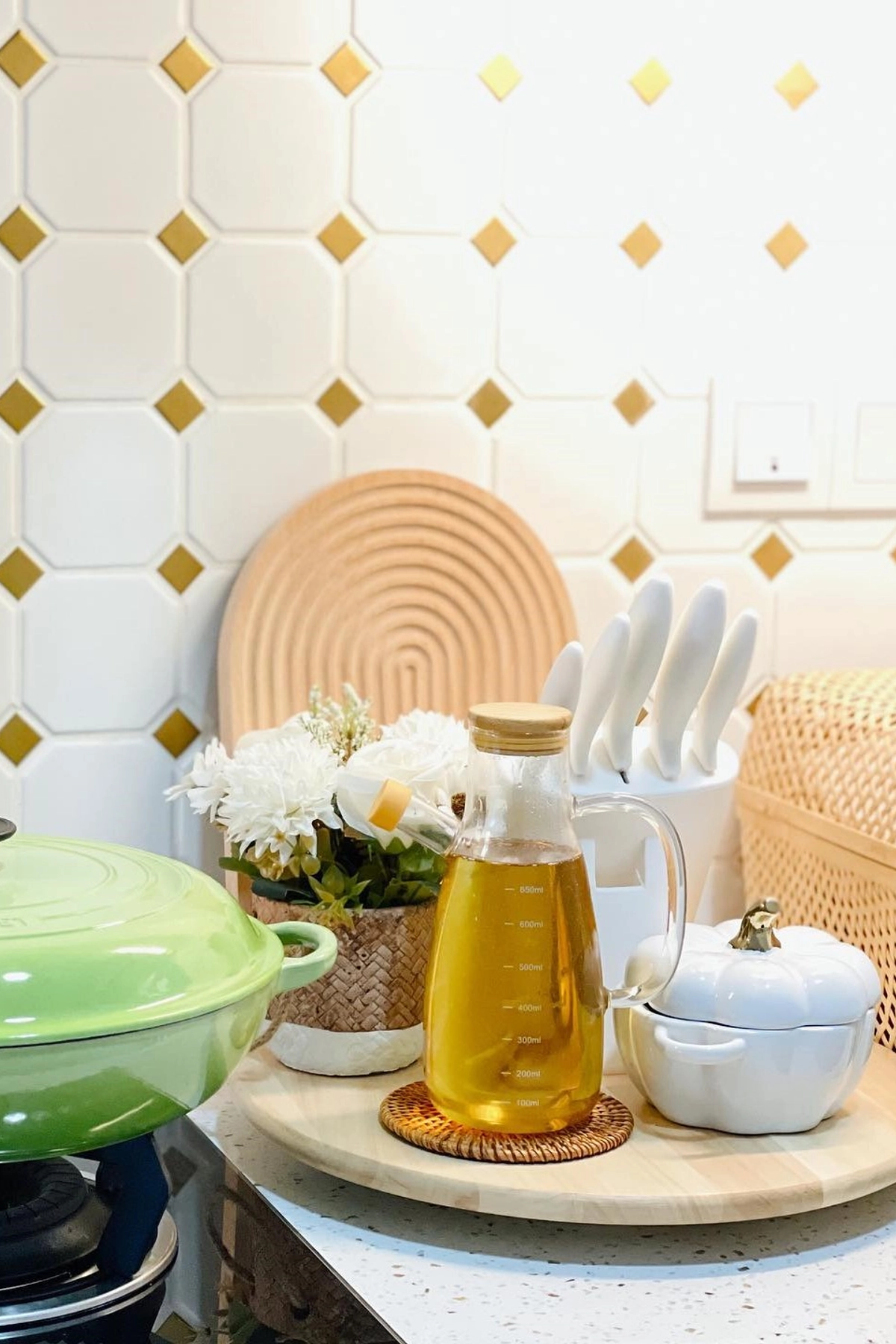 Green pot on stovetop, honey jar, white flowers in basket, decorative board against a white and gold tiled wall.