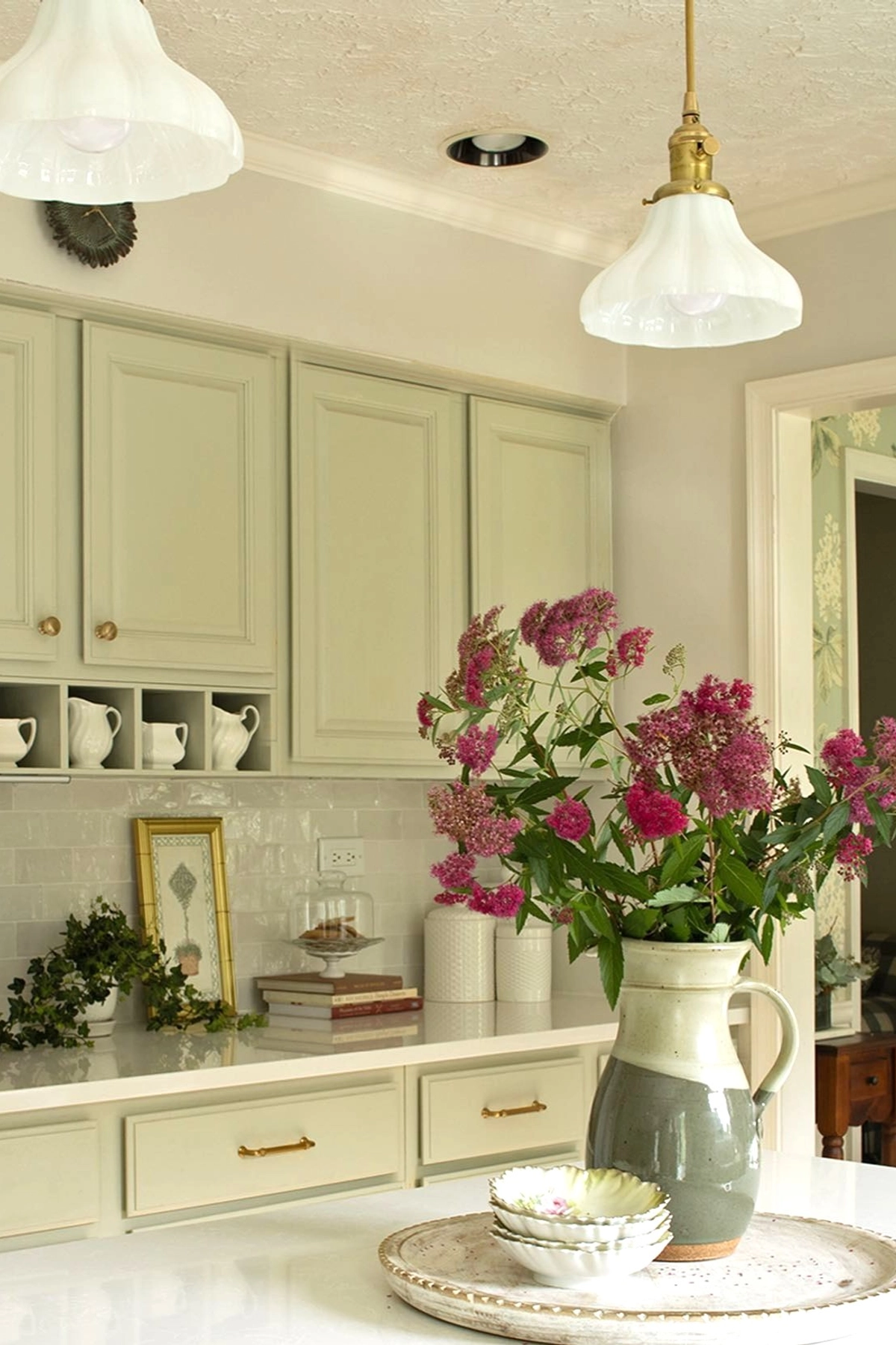 Light kitchen with pale green cabinets, pink flowers in a large vase, white ceramic dishes, and framed art on the countertop.