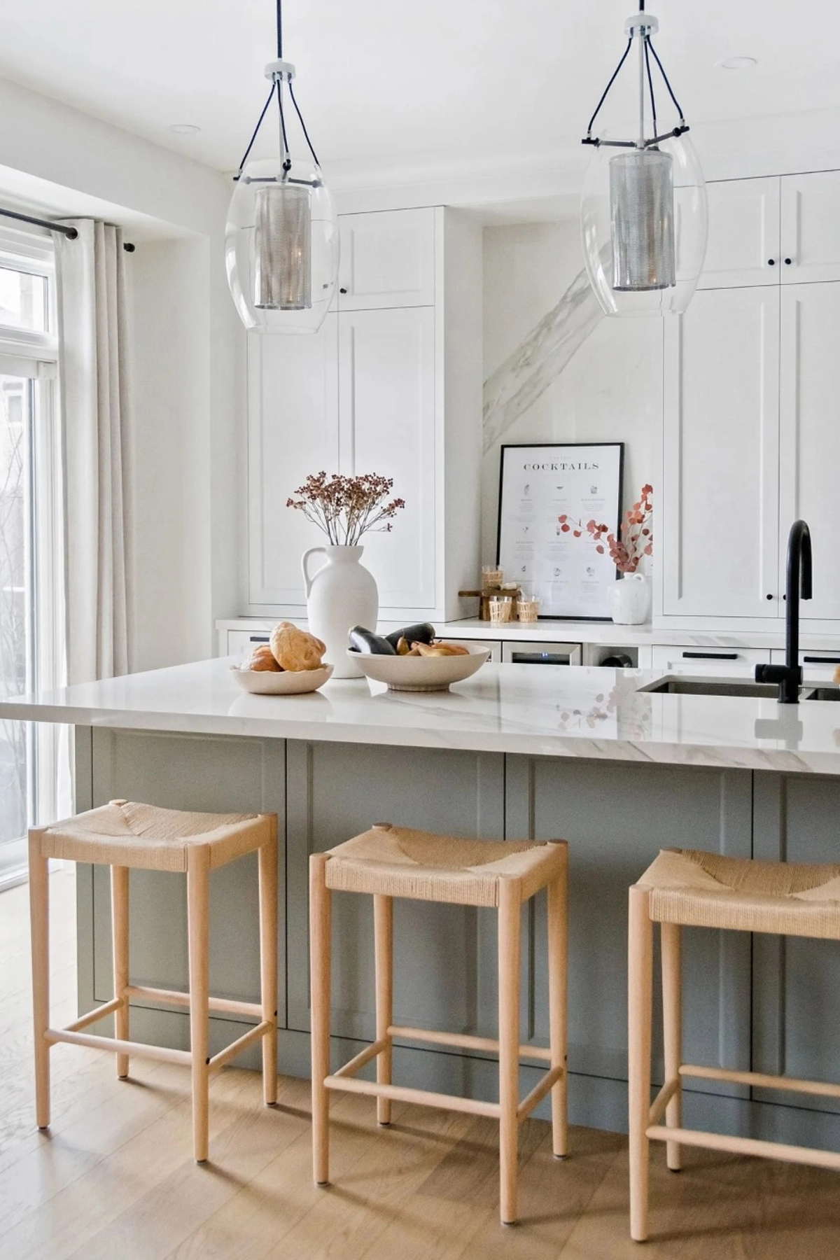 Bright kitchen with white countertops, light wood stools, pendant lights, and decorative bowls with bread.