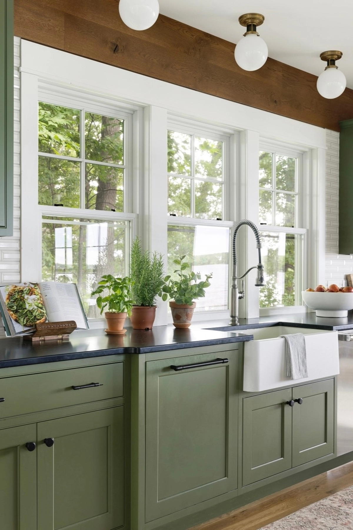 Green kitchen cabinets under large windows with potted herbs, a recipe book stand, and a bowl of fruit on the countertop.