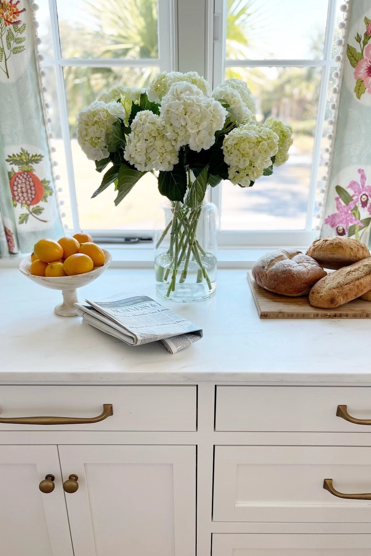 White kitchen countertop with a vase of white flowers, bowl of lemons, newspaper, and bread by a window.