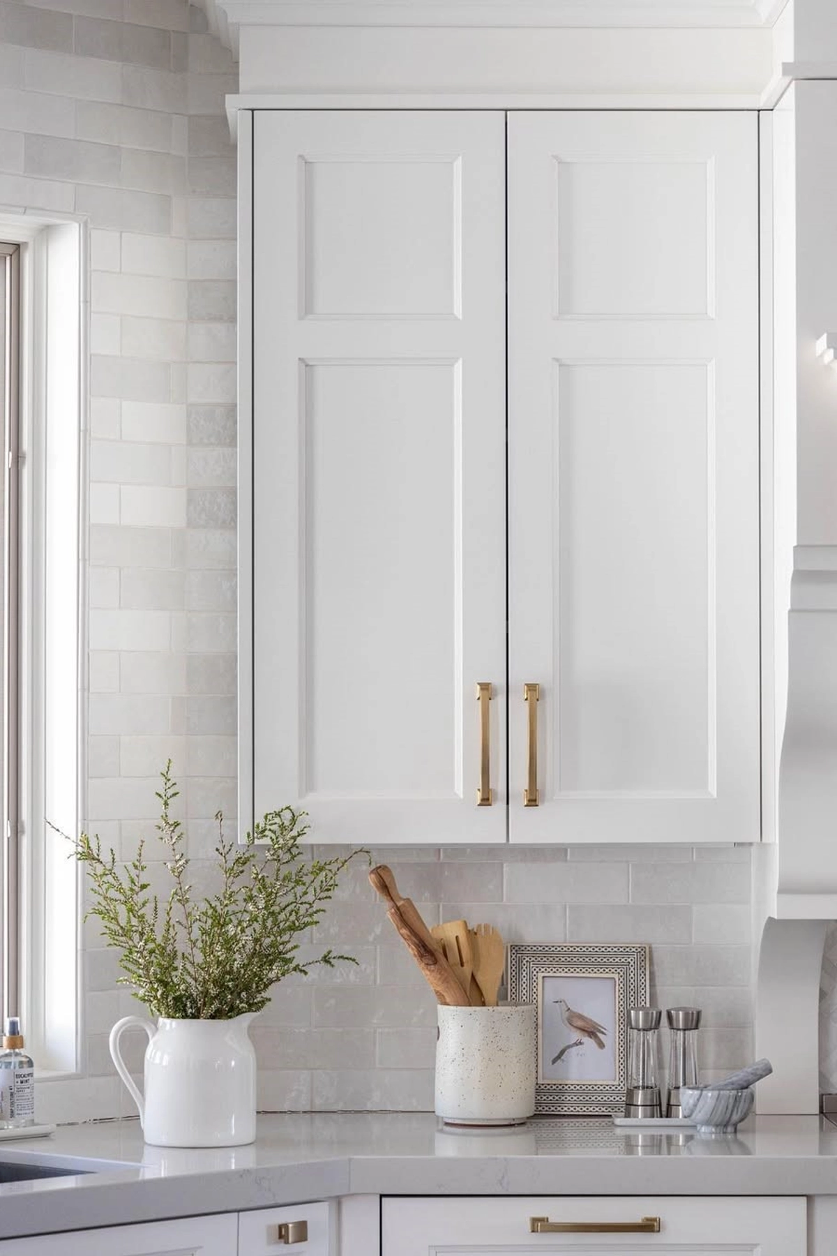 Kitchen counter with white cabinets, a white vase with greenery, a mortar and pestle, utensils in a ceramic holder, and a small picture frame.