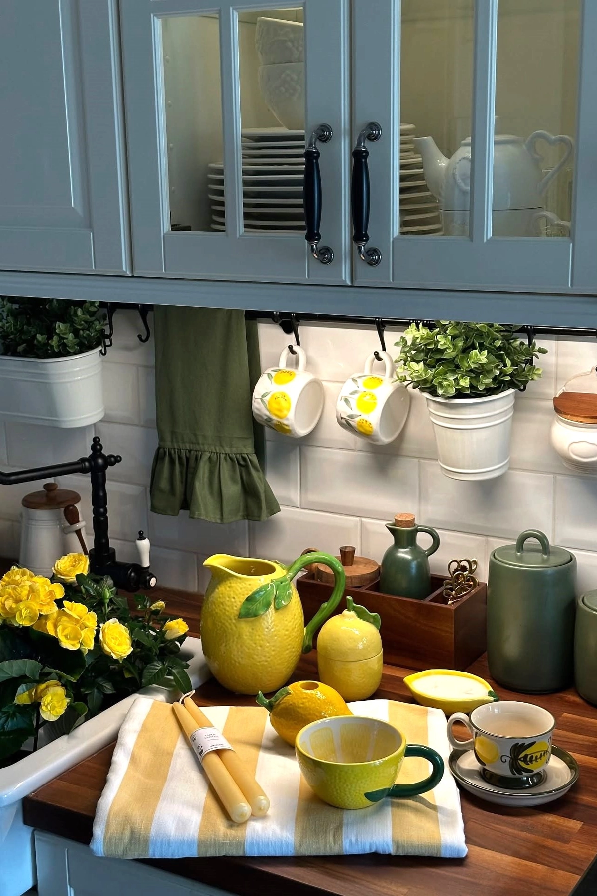 Kitchen countertop with lemon-themed pitcher, mugs, yellow flowers, green towel, and hanging plants under cabinets.