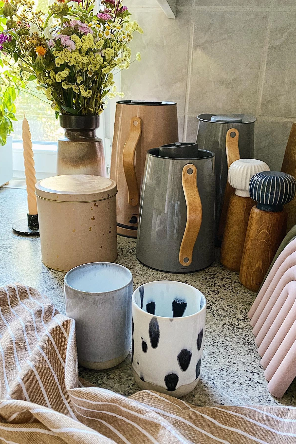 Kitchen countertop with patterned mugs, ceramic jars, wildflowers in a vase, wooden accents, and striped cloth.