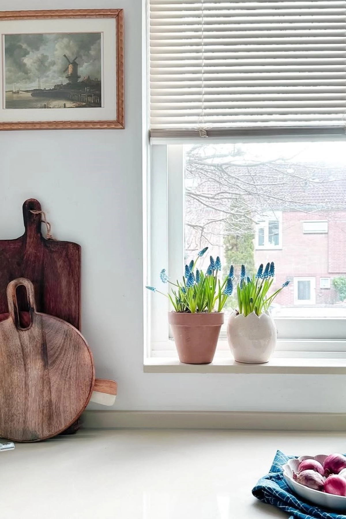 Kitchen countertop with cutting boards, potted blue flowers on the windowsill, framed art, and onions in a bowl.