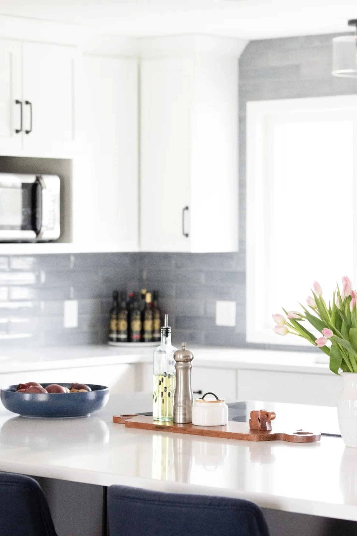 White kitchen countertop with pink tulips in a vase, olive oil bottle, pepper mill, and sunlight streaming through a window.