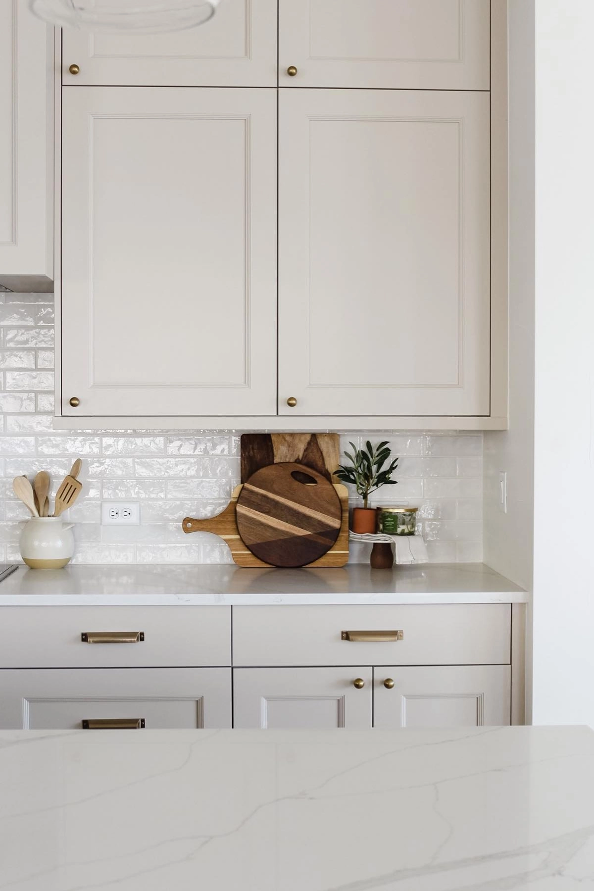 Neutral kitchen with white cabinets, gold handles, wooden cutting boards, utensils in a jar, and small plant on the countertop.