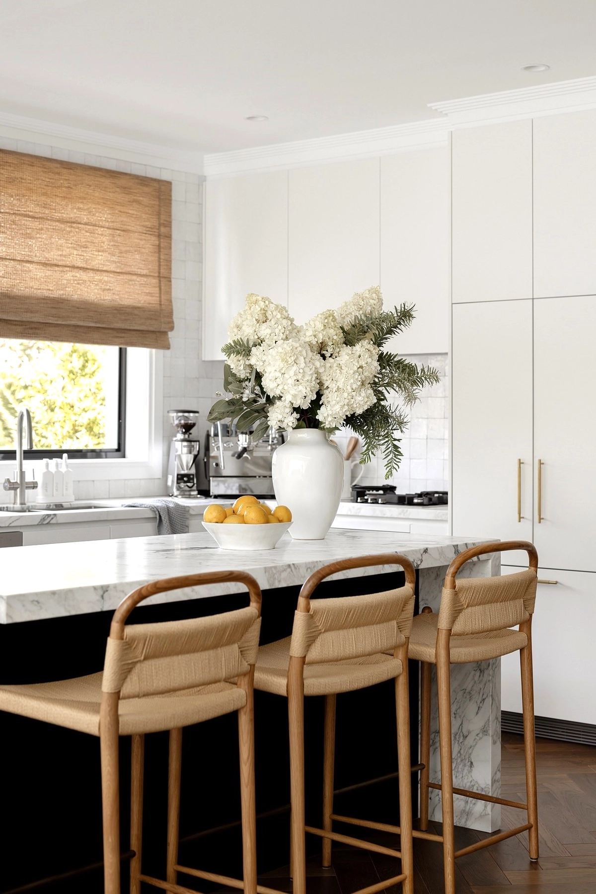Kitchen with white cabinets, large floral vase, lemons in a bowl on marble countertop, and wooden stools.