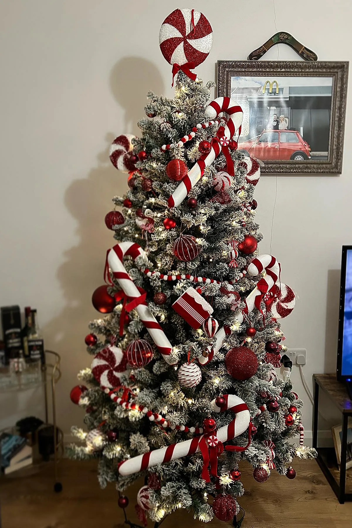 Frosted Christmas tree decorated with large red and white striped candy cane ornaments, peppermint swirl toppers, red baubles, and white and red garlands set in a cozy living room with wooden flooring.