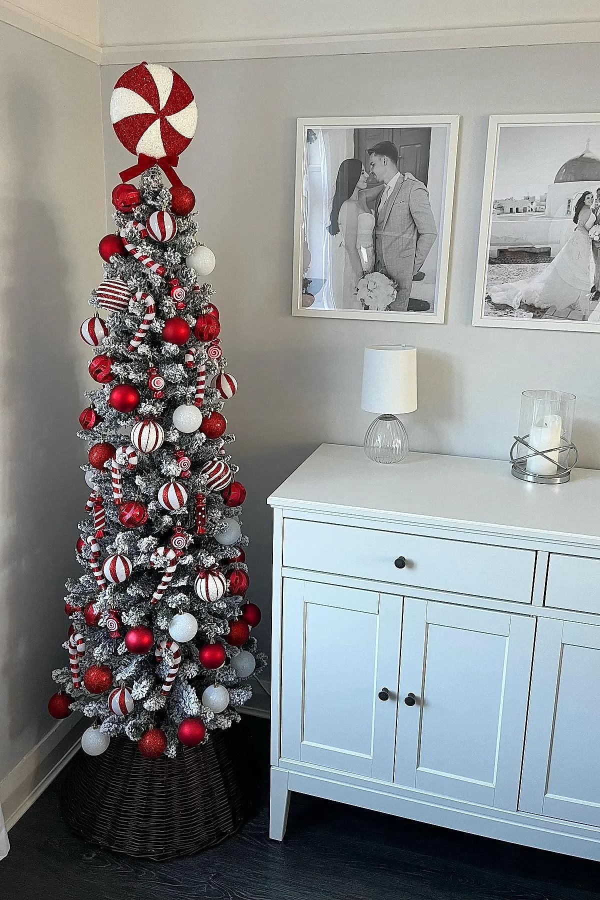 Frosted Christmas tree decorated with candy cane themed ornaments including striped balls, candy cane shaped decorations, and a large peppermint swirl topper next to a white cabinet with black knobs beneath black and white wedding photos.