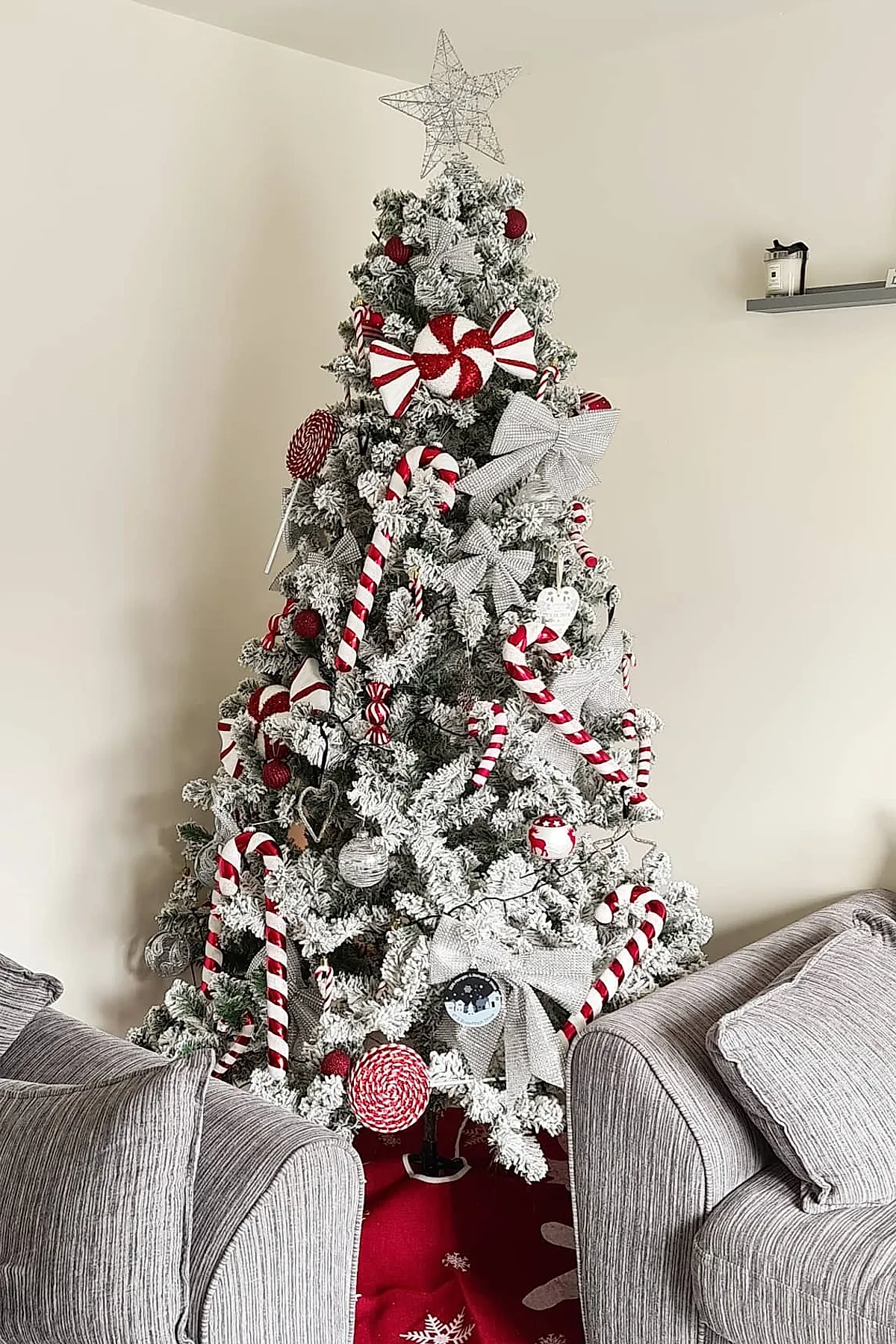 Flocked Christmas tree decorated with red and white candy cane ornaments, oversized silver bows, peppermint swirl decorations, and a wire star topper placed between two gray fabric sofas in a neutral living room.