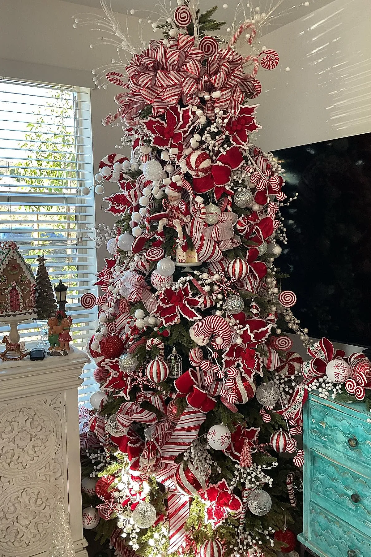 A lush Christmas tree decorated in a candy cane christmas tree theme featuring large red and white striped ribbons, red velvet poinsettia flowers with white trim, red and white peppermint swirl ornaments, white berry branches, silver baubles, and candy cane decorations. The tree stands next to a turquoise distressed dresser and near a white carved mantel with holiday figurines.