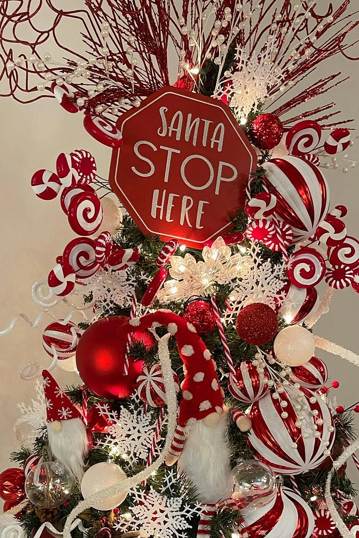 Christmas tree decorated with a red and white candy cane theme including large peppermint swirl ornaments, red baubles, white snowflakes, glittery branches, and a red sign reading Santa Stop Here at the top.