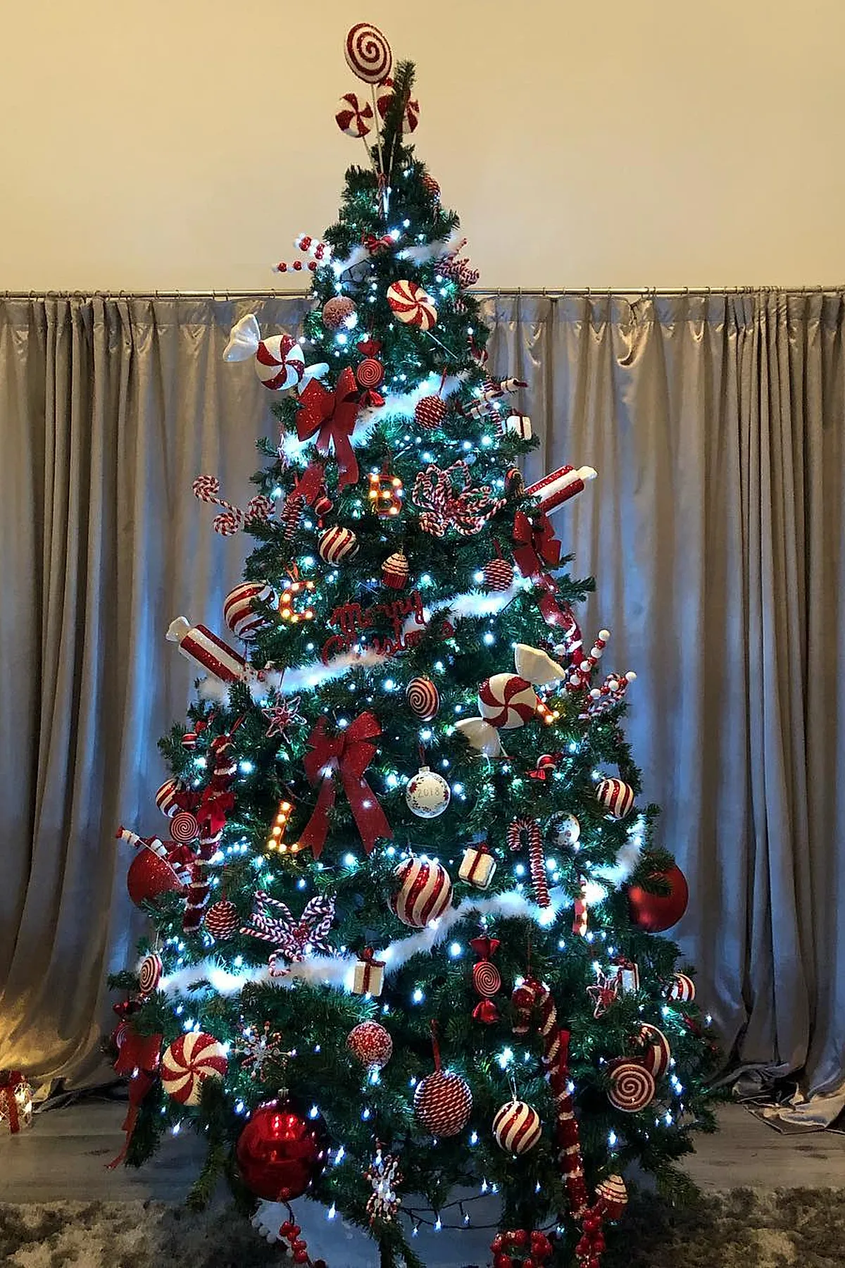 Christmas tree decorated with a candy cane christmas tree theme featuring red and white peppermint swirl ornaments, large red bows, candy cane shaped decorations, white garlands, and warm white string lights in front of beige curtains.