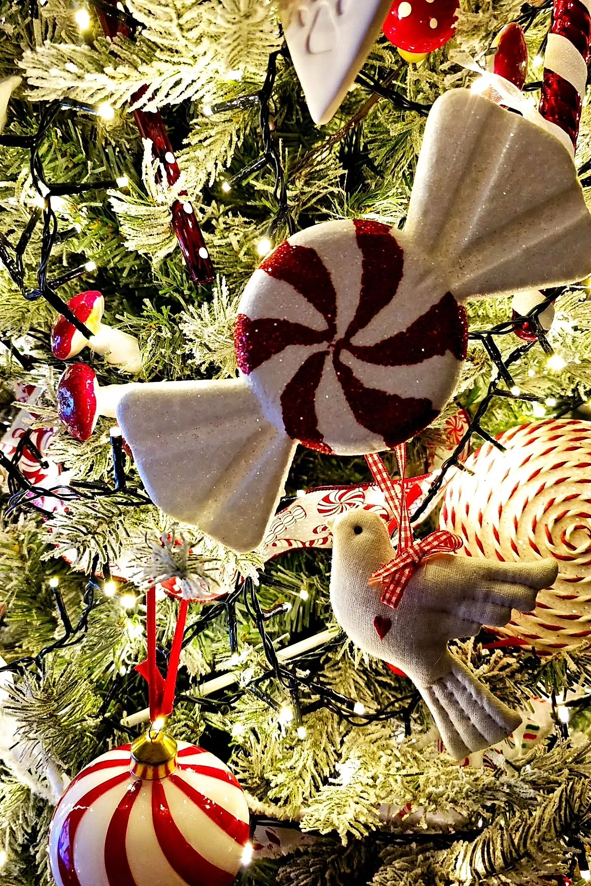 Close up of a candy cane christmas tree theme featuring glittery red and white peppermint swirl ornaments, a white fabric bird with a red heart and gingham ribbon, small red and white mushroom decorations, and warm white string lights on green frosted pine branches.