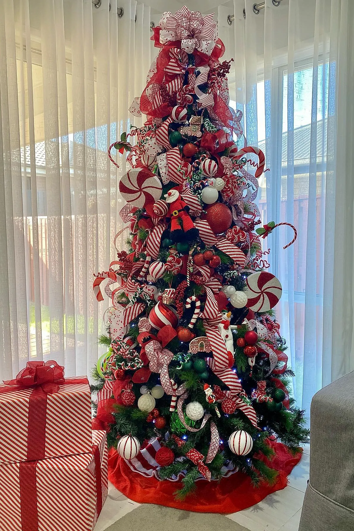 Lush Christmas tree decorated in a candy cane christmas tree theme featuring large red and white striped ribbons, oversized peppermint swirl ornaments, red, white, and green baubles, candy cane shaped decorations, plush Santa figures, and red berries placed near sheer white curtains with wrapped gifts in matching red and white striped paper.