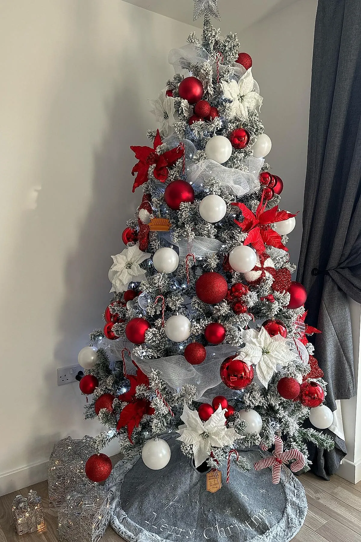 Frosted Christmas tree decorated in a candy cane christmas tree theme with red and white baubles, large white poinsettias, silver mesh ribbon, candy cane ornaments, and a silver star topper next to grey curtains on wooden floor.