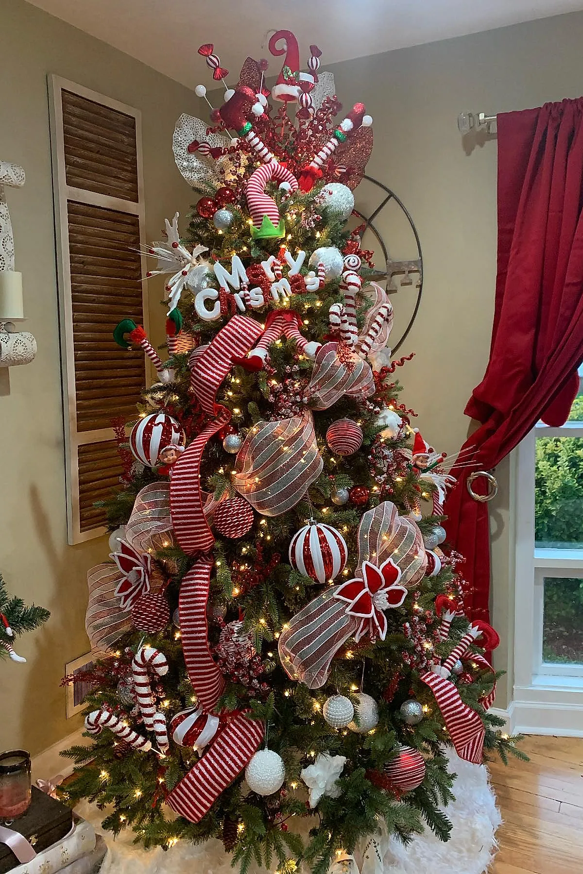 Christmas tree decorated in a candy cane christmas tree theme featuring red and white striped ribbons, large red and white baubles, glittery mesh ribbons, elf hat ornaments, candy cane shaped decorations, white berry clusters, and warm string lights in a cozy living room corner near a window with red curtains.