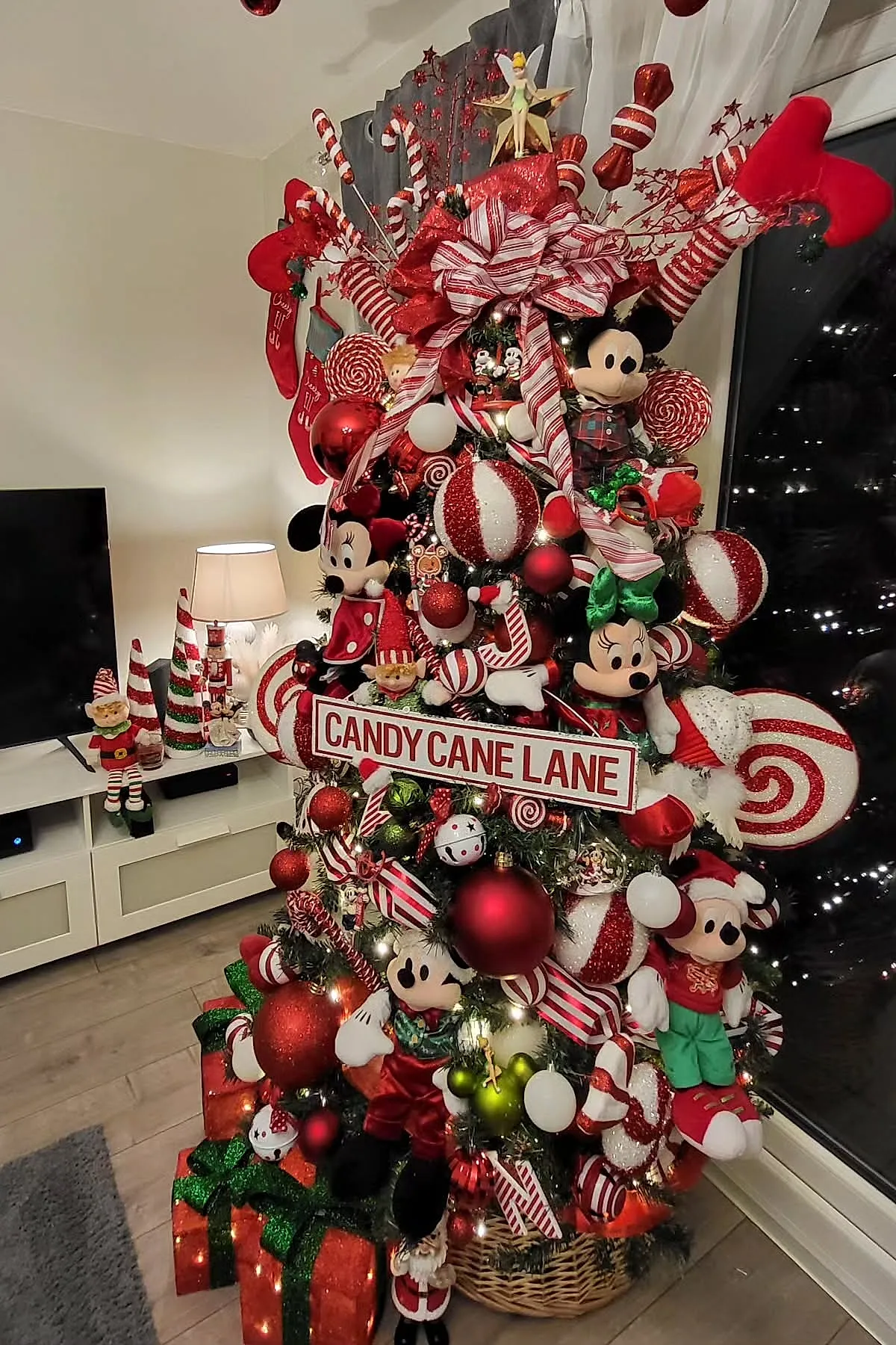 A candy cane christmas tree theme decorated with red and white striped ribbons, oversized peppermint swirl ornaments, red baubles, Mickey and Minnie Mouse plush toys dressed in holiday attire, elf figurines, and a Candy Cane Lane sign. The tree stands next to a window with sheer curtains in a modern living room.
