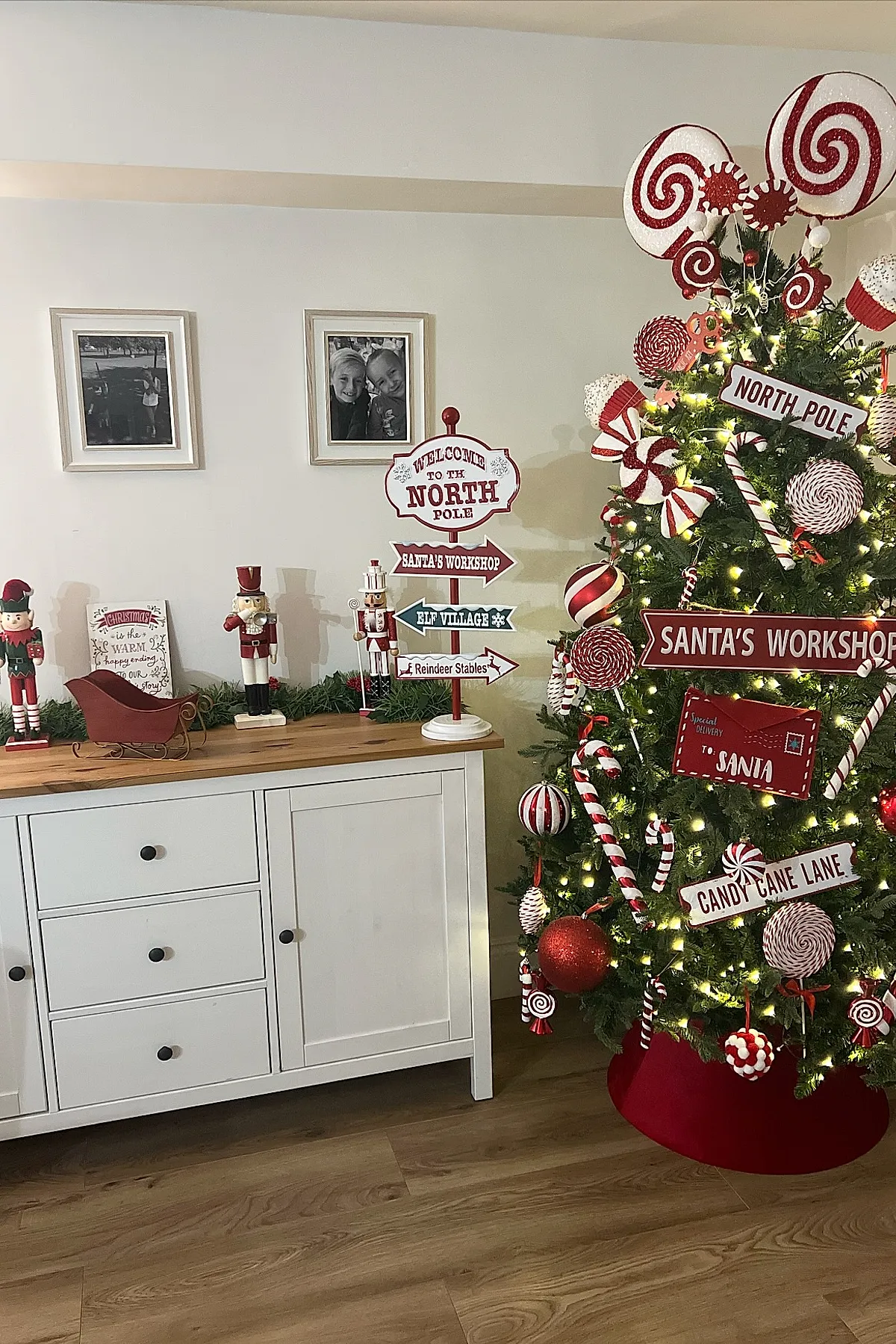 Christmas tree decorated with a candy cane christmas tree theme featuring red and white peppermint swirl ornaments, candy canes, large lollipops, and directional signs labeled North Pole, Santa's Workshop, and Candy Cane Lane, standing next to a white cabinet topped with nutcracker figurines and a small red sleigh.