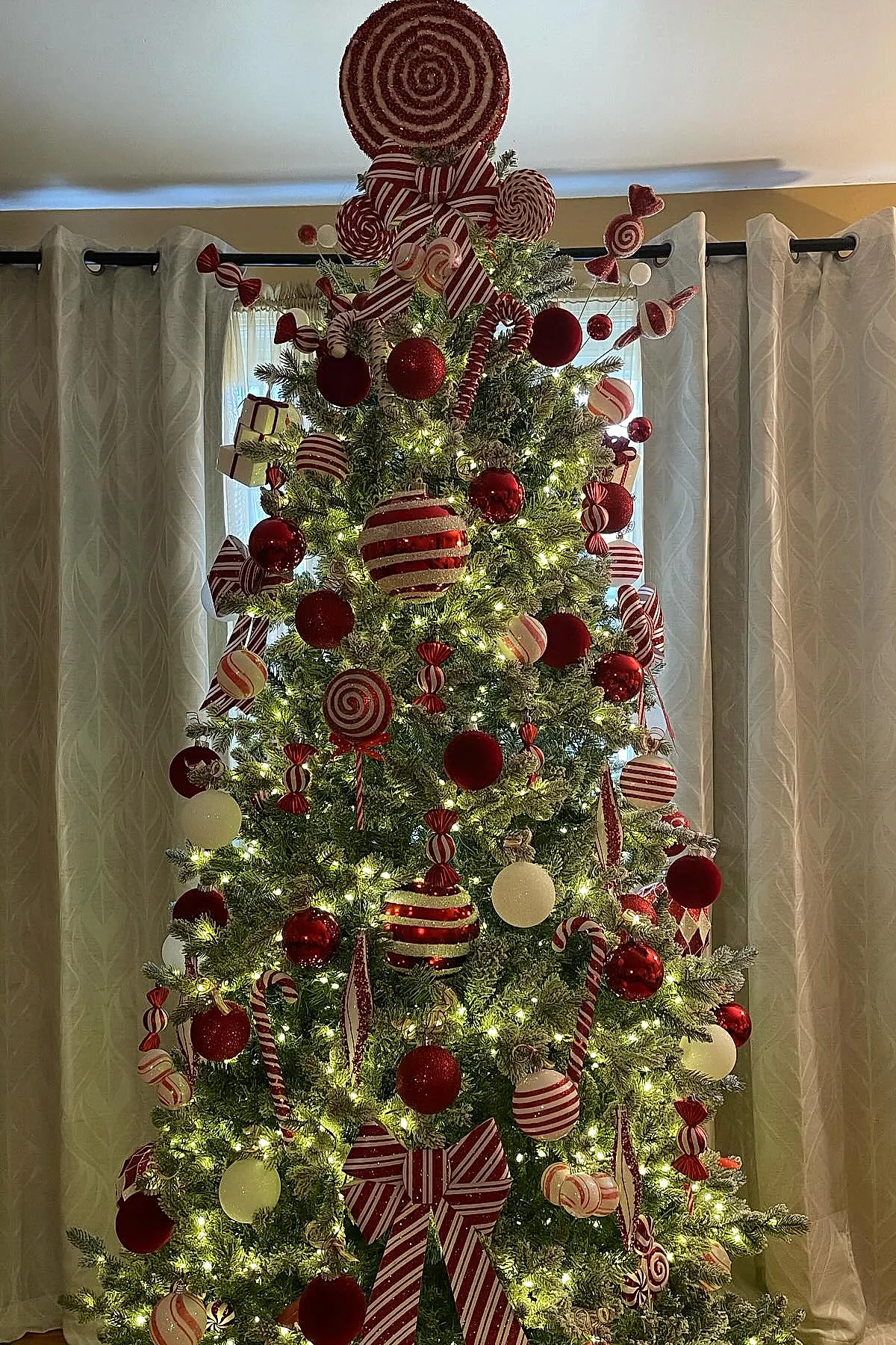 Christmas tree decorated in a candy cane christmas tree theme with red and white striped bows, large lollipop ornaments, candy cane decorations, and red and white baubles lit by warm string lights in front of beige curtains.