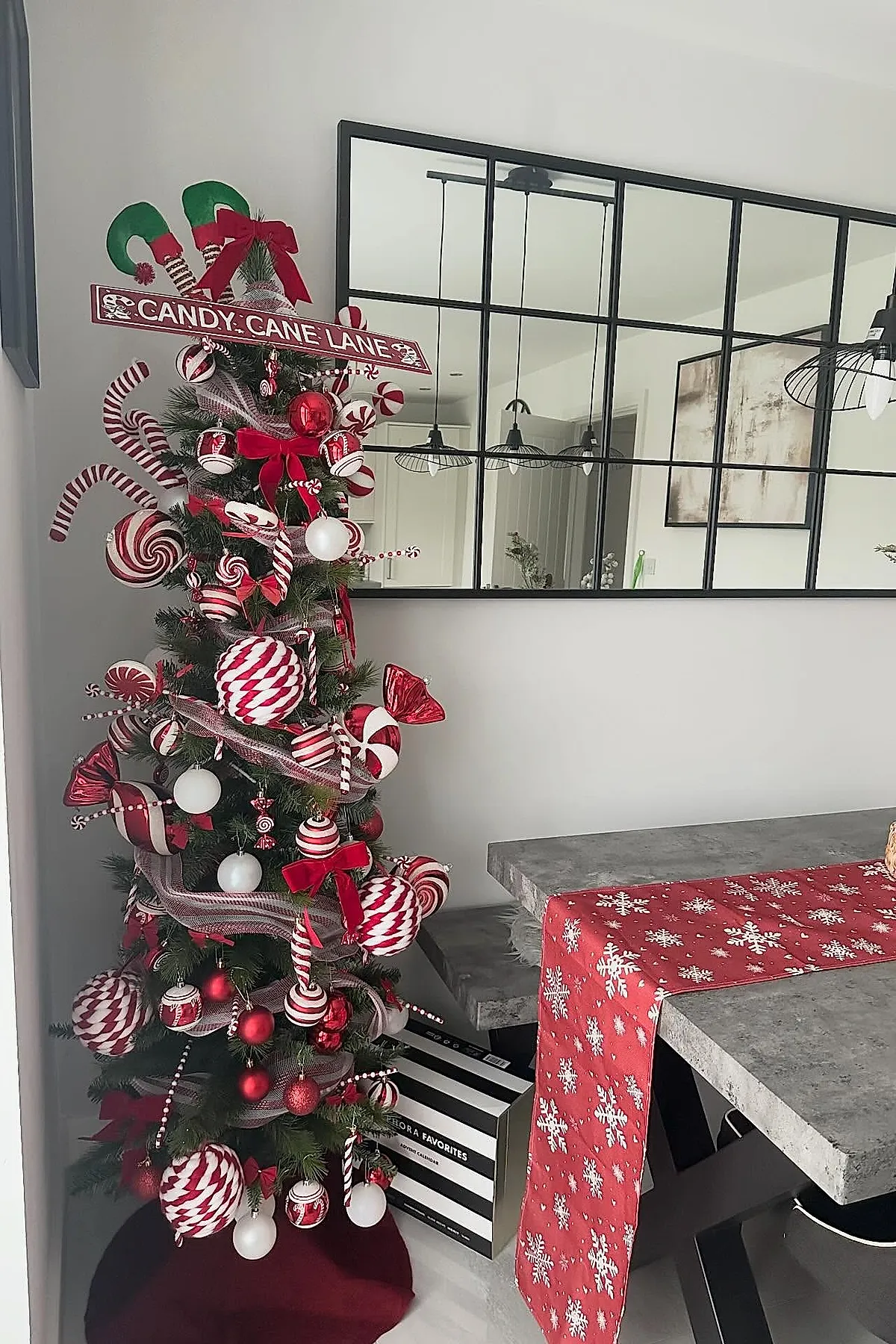 Tall Christmas tree decorated with a candy cane christmas tree theme featuring large red and white swirled ornaments, oversized candy cane decorations, red bows, a Candy Cane Lane sign topper with green elf shoe accents, white round baubles, and ribbon garlands beside a concrete dining table with a red snowflake runner.