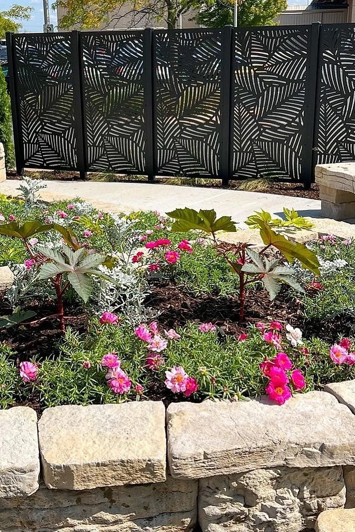 Black metal privacy screen with fern patterns behind a stone planter filled with colorful flowers.