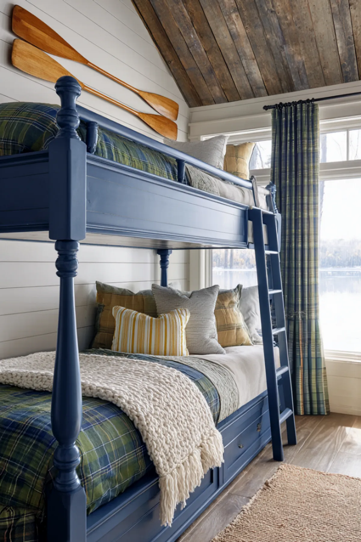 Blue built-in bunk beds with plaid bedding, rustic paddles on shiplap walls, and large windows offering natural light.